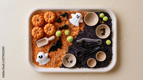 Halloween sensory bin with orange pumpkins, white ghosts, black bats, colorful rice, wooden scoops and bowls for playful autumn learning