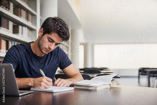 A young man is studying and writing in a notebook at a table in a bright, modern library or study area.