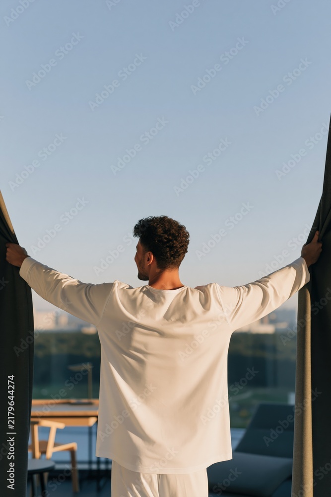 Obraz premium Rear view man opening curtains on a balcony at sunrise with urban skyline for interior background and morning routine banner