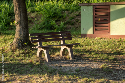 Hölzerne Sitzbank mit Betonfüßen im Seitenlicht der tiefstehenden Sonne vor ländlichem Trafohäuschen in Neckartenzlingen