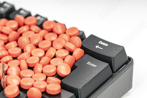 Close-up of numerous orange pills scattered over a black computer keyboard next to the enter and delete keys, representing health, medication and online pharmacy risks