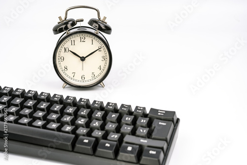 Vintage twin-bell alarm clock positioned behind a modern black computer keyboard on white background, symbolizing time management, deadlines, and the consequences of pressing enter in digital tasks
