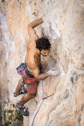 Shirtless climber ascends a steep rock face. His hands grip the stone securely. He shows intense determination and effort. Climbing gear and rope secure his position