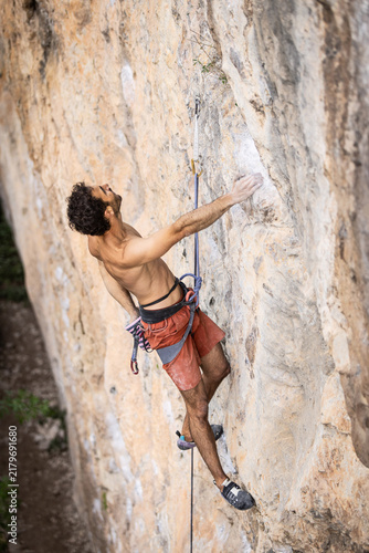 Shirtless climber grips a rocky cliff face. He wears orange shorts and a climbing harness. His arms strain against the stone wall. His focused expression shows determination. Perfect for adventure