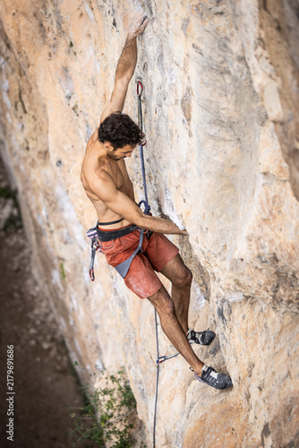 Shirtless man climbs a steep rock face. He grips the stone with focused effort. His red shorts and harness stand out against the rock. Natural terrain shows real climbing challenge