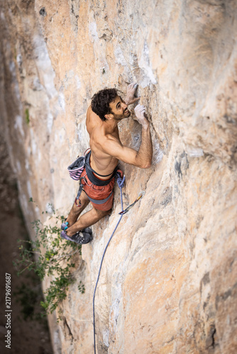 Shirtless man climbs a rugged rock face. His hands grip the stone firmly. Blue climbing ropes secure his position. His expression shows intense focus. This captures outdoor adventure energy