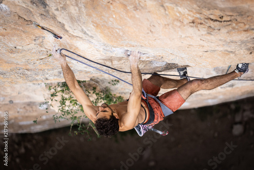 Climber grips the rock face with focused determination. His orange shorts and harness show climbing gear. He uses ropes for safety during ascent. The textured rock wall is light brown
