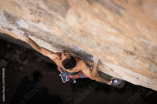 Shirtless climber grips rocky overhang. Chalk dust marks his hands. He focuses intensely upward. Dark rock face contrasts his bright gear. High angle shows his strong balance