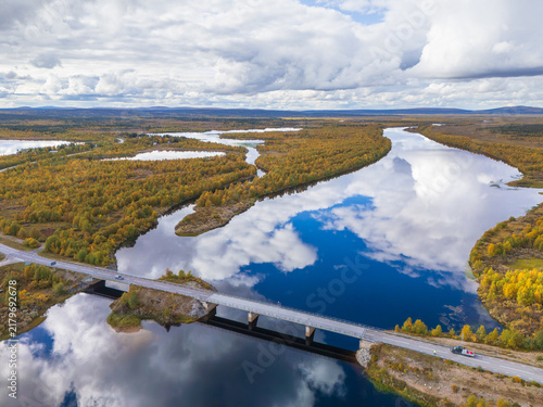 Принти на полотні Aerial view of a bridge crossing a mirror-like river reflecting clouds surrounde