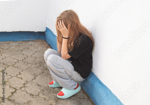фотографія A teenage girl is sitting on her heels near a white wall, clutching her head in