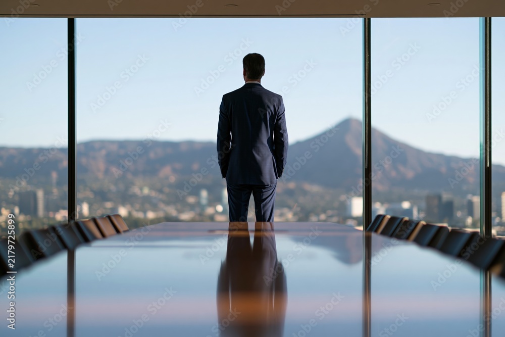 Naklejka premium Business executive standing at a boardroom table by floor to ceiling windows with mountain cityscape beyond for corporate office background and leadership banner