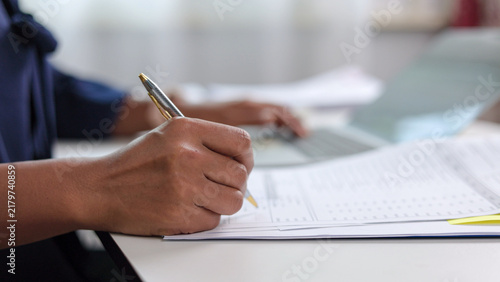 Businesswoman hand holding pen and writing on paper document at office desk. Female accountant signing agreement contract and checking financial report with laptop computer.
