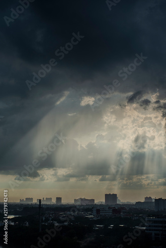 Canvas Print Dramatic crepuscular rays breaking through dark storm clouds above the silhouetted skyline of Ho Chi Minh City creating a powerful and moody atmospheric cityscape before a tropical storm