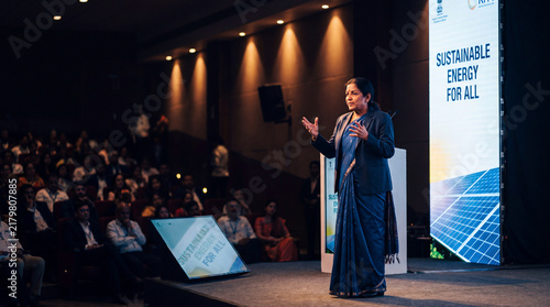 Empowering Minds: A charismatic speaker commands the stage, addressing an engaged audience in a modern auditorium, illuminated by a backdrop highlighting sustainable energy solutions.