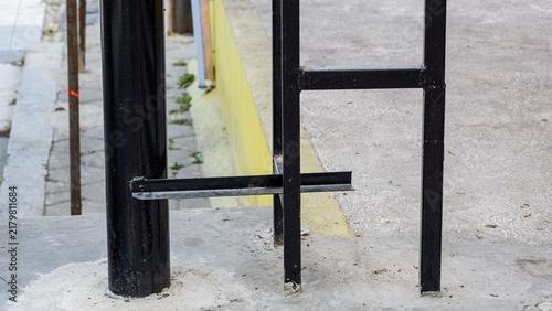 Obraz na płótnie Black iron ladder structure anchored into a concrete ground surface alongside a vertical metal pole on an urban sidewalk