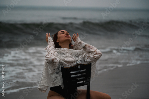 Obraz na płótnie Model poses on a chair near the ocean while waves wash ashore during sunset
