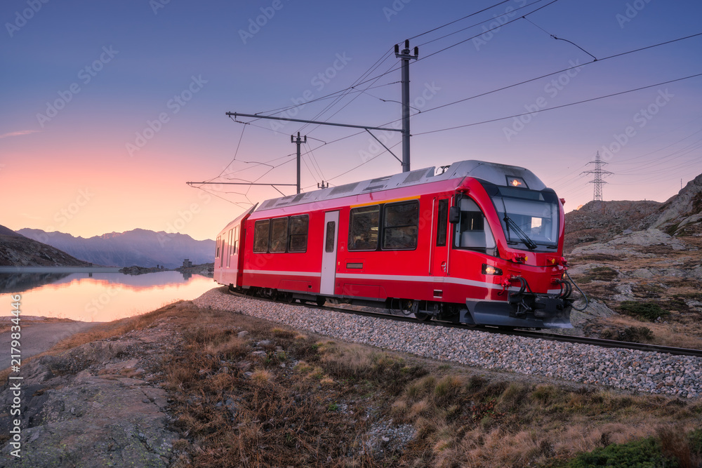 Fototapeta premium Bernina Express train near Lake Bianco in Switzerland at sunset, purple sky, clear reflections, mountain silhouettes and alpine terrain in evening light in the Swiss Alps. Red train at dusk. Transport