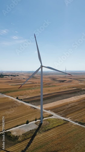 Renewable energy and sustainability concept, aerial vertical drone view flying to the left of a single wind turbine standing in a rural agricultural field under a clear blue sky