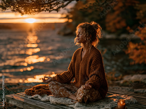 Woman sits cross‑legged meditating on a wooden dock beside calm water, warm sunset golden light glowing over peaceful lakeside scenery.