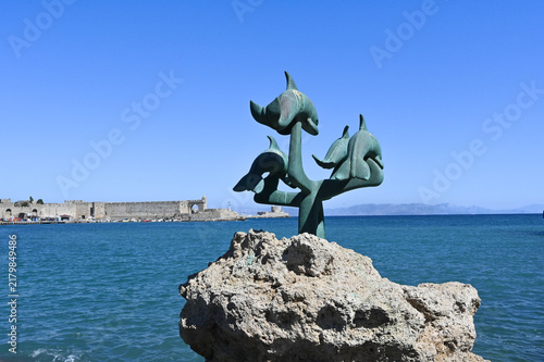 Bronze dolphin sculpture by the sea with fortress in background - Old Town Rhodes, Greece.