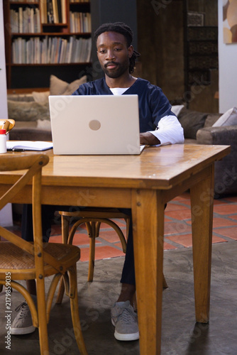 Silver laptop is sitting open on wooden dining table with notebook, pen holder, bookshelf behind
