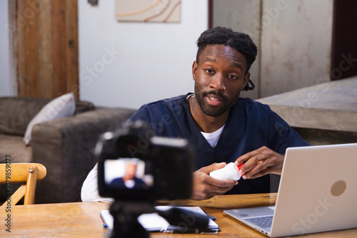 African American man wearing scrubs at home showing white bottle on table with laptop and camera
