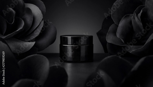 Black cosmetic cream jar surrounded by dark flowers on a black background