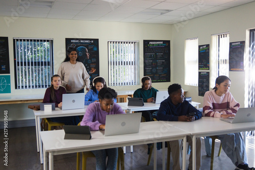 Diverse students with Indian teacher supervising typing on laptops and tablets in classroom