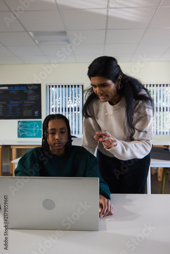 Indian tutor leaning over guiding African American Youth student using silver laptop in classroom