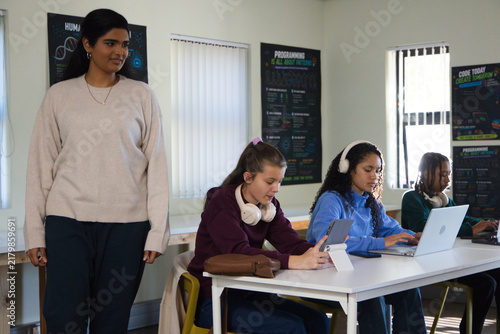 Instructor with teenage students standing and working at class table using laptops tablet headsets