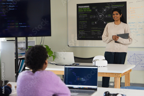 Indian teacher standing with tablet in lab while African youth student coding on laptop, copy space