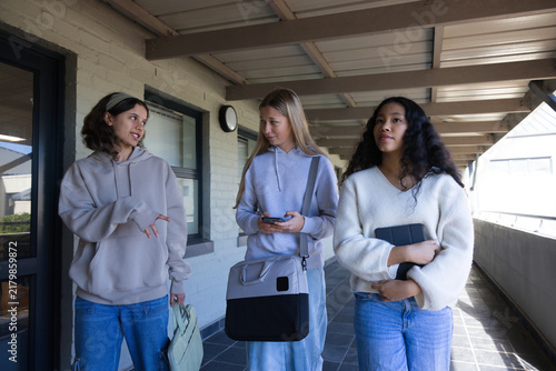 Diverse teenage females walking covered corridor by painted brick wall, holding smartphone, tablet