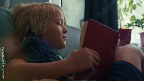 A young blond boy sits in a comfortable armchair by the window and reads a book at home. The cozy afternoon scene captures childhood, education, relaxation, and peaceful indoor leisure.