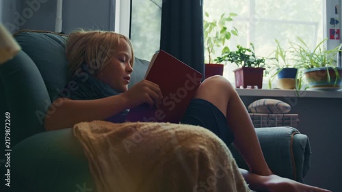 Little boy with blond hair enjoys a quiet afternoon reading a book in a cozy room. The footage represents learning, concentration, literature, and a comfortable home lifestyle.