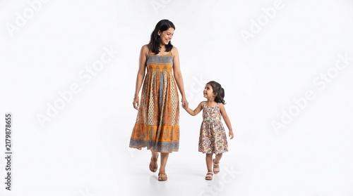 A happy mother and her young daughter walking hand in hand, wearing matching ethnic floral print dresses, isolated on white background