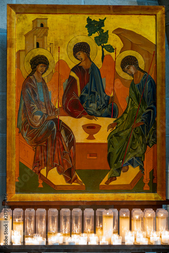 Фотография An icon panel depicting three angels sits above lit votive candles displayed for devotional prayer
