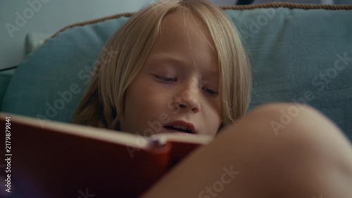 Young child relaxes in an armchair while reading a book near a bright window. The peaceful indoor setting creates an atmosphere of curiosity, knowledge, concentration, and relaxation.