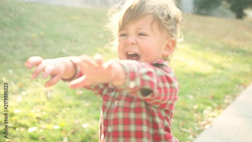 Toddler in a red plaid shirt laughing excitedly while walking on a leaf-covered sidewalk
