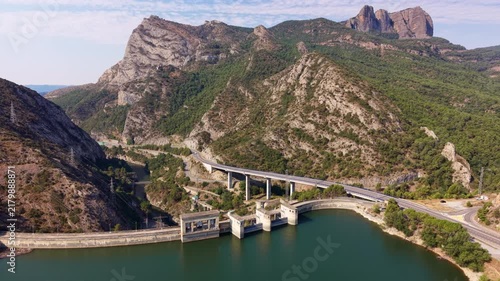 Aerial view of a concrete dam and reservoir nestled between rugged rocky mountains and forested slopes, with a highway viaduct crossing above