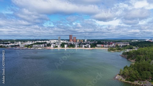 Panoramic view of Vuosaari district in Helsinki, Finland, showing a sandy bay along the Baltic Sea shoreline with residential buildings, green forest and cloudy sky