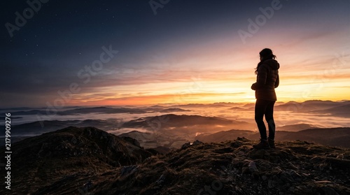 Woman standing on mountain peak watching sunrise over foggy valley