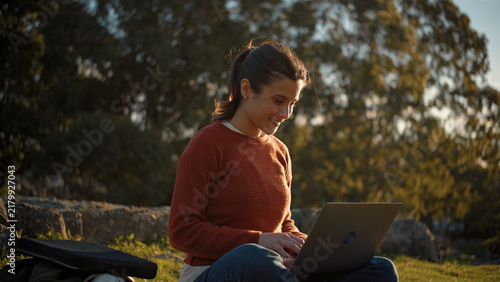 Young Latina woman sitting on the grass in a park using her laptop at sunset, with trees in the background, highlighting productivity, nature, and a peaceful outdoor lifestyle.