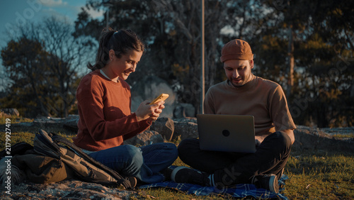 Young Latino heterosexual couple sitting in a park using technology together, highlighting digital connection, companionship, communication, and a modern outdoor lifestyle.