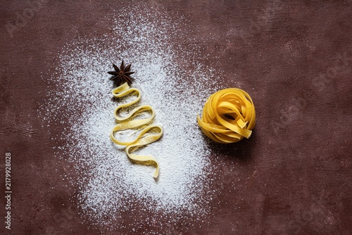 Raw yellow italian pasta (nests) pappardelle, fettuccine or tagliatelle in the shape of a Christmas tree on a brown background, close up. Egg homemade noodles cooking process,festive mood.