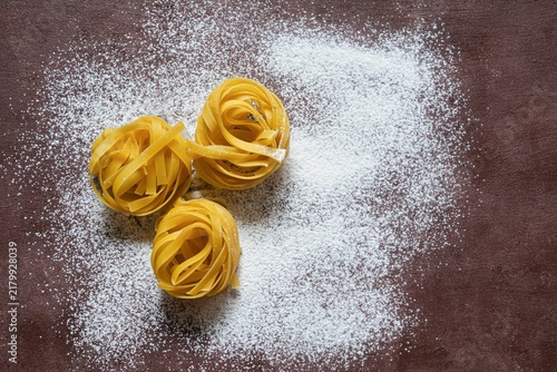 Raw yellow italian pasta (nests) pappardelle, fettuccine or tagliatelle on a brown background, close up. Egg homemade noodles cooking process, festive mood.