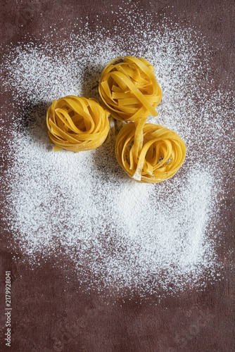 Raw yellow italian pasta (nests) pappardelle, fettuccine or tagliatelle on a brown background, close up. Egg homemade noodles cooking process, festive mood.