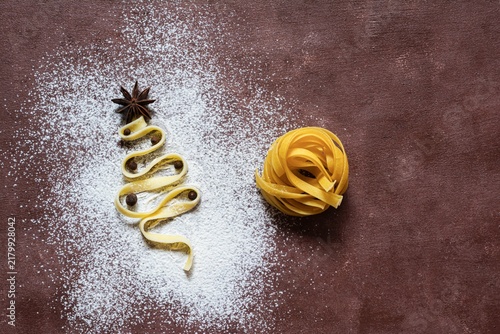 Raw yellow italian pasta (nests) pappardelle, fettuccine or tagliatelle in the shape of a Christmas tree on a brown background, close up. Egg homemade noodles cooking process,festive mood.