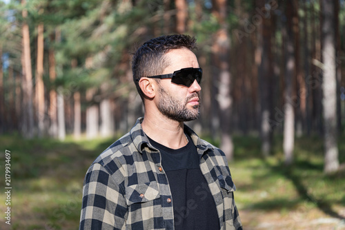 Vertical outdoor portrait of a confident bearded man wearing tactical sunglasses and a plaid shirt standing in a sunny pine forest during daytime.