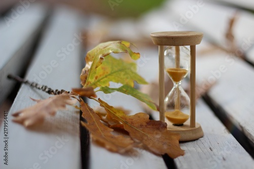 Hourglasses and yellow oak leaves on a white wooden bench — the concept of the changing seasons, a time for rest and walks, leaf fall, the autumn season of life