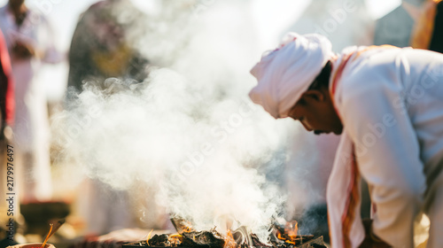 Close-up of thick incense smoke rising during a Hindu Antyesti ceremony while a male mourner in traditional white clothing bows beside the sacred fire, relatives softly blurred in